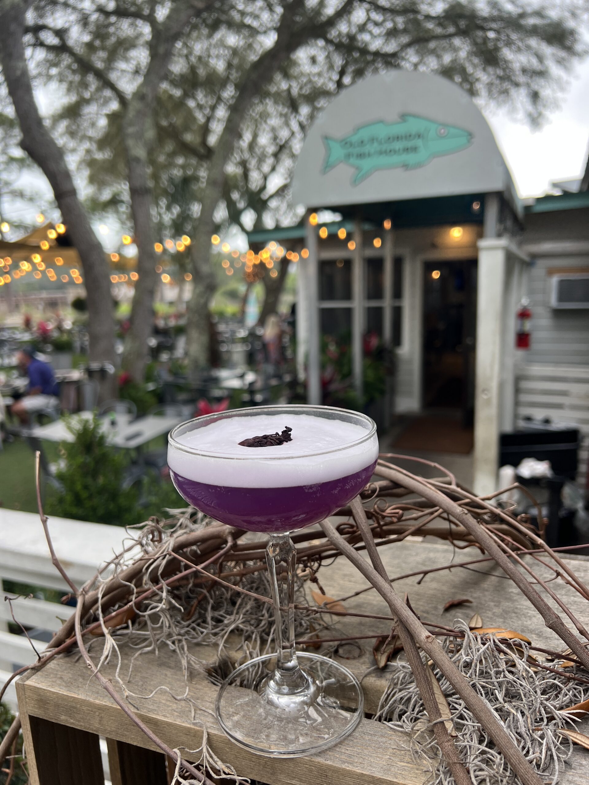 Signature purple craft cocktail served in a coupe glass on the patio at Old Florida Fish House with the restaurant entrance in the background.