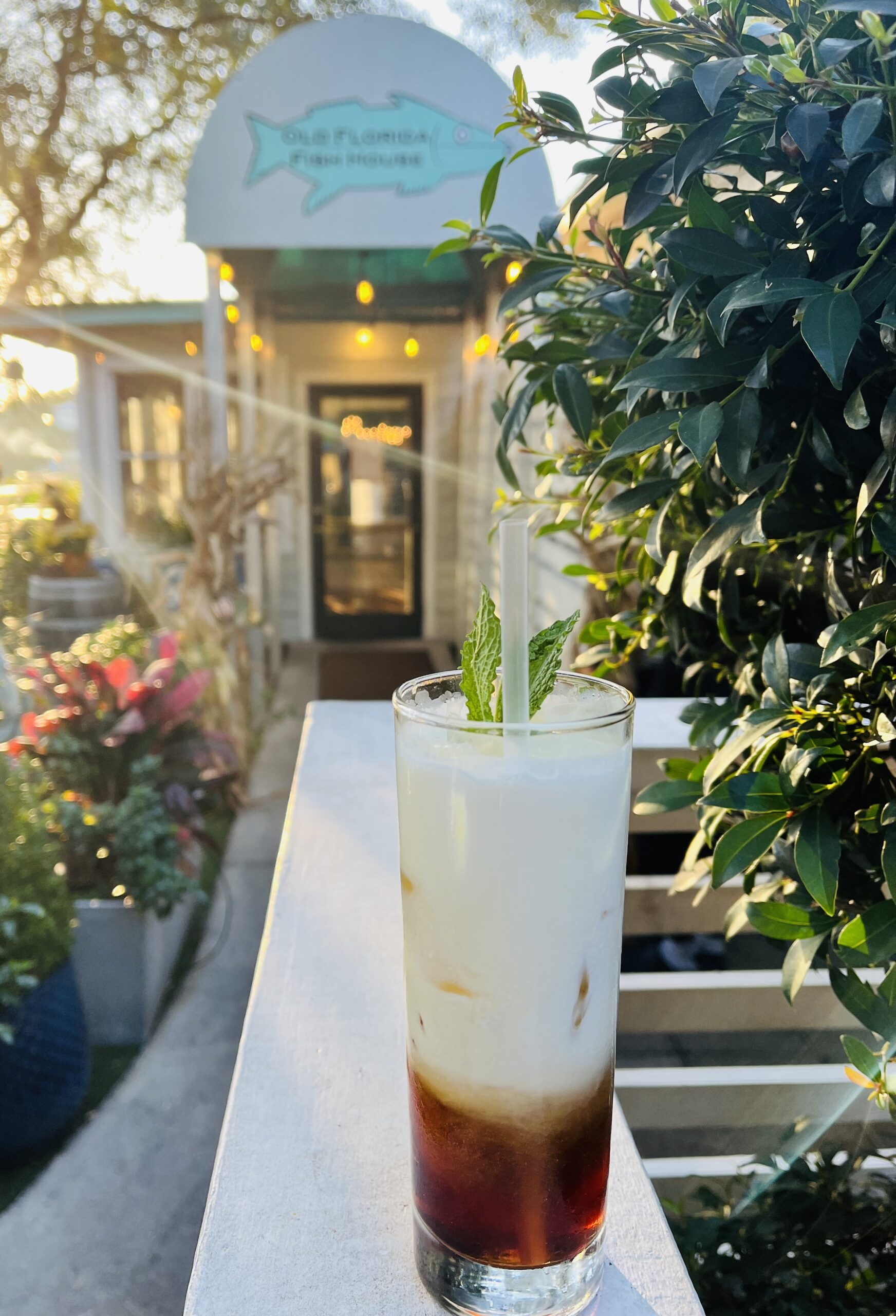 Creamy layered cocktail with dark coffee liqueur and frothy white top, garnished with fresh mint, served in a tall glass on the patio at Old Florida Fish House with the restaurant entrance softly blurred in the background.