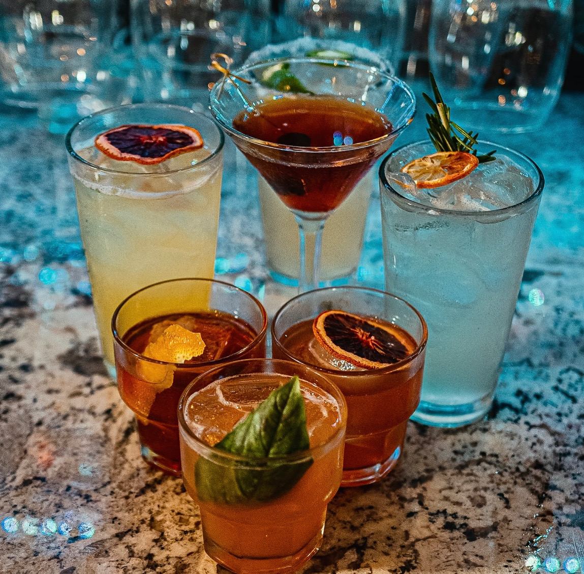 Assortment of handcrafted cocktails arranged on a granite bar, including martinis, citrus highballs, and spirit-forward drinks garnished with dried blood orange slices, rosemary, citrus peel, cherries, and fresh basil.