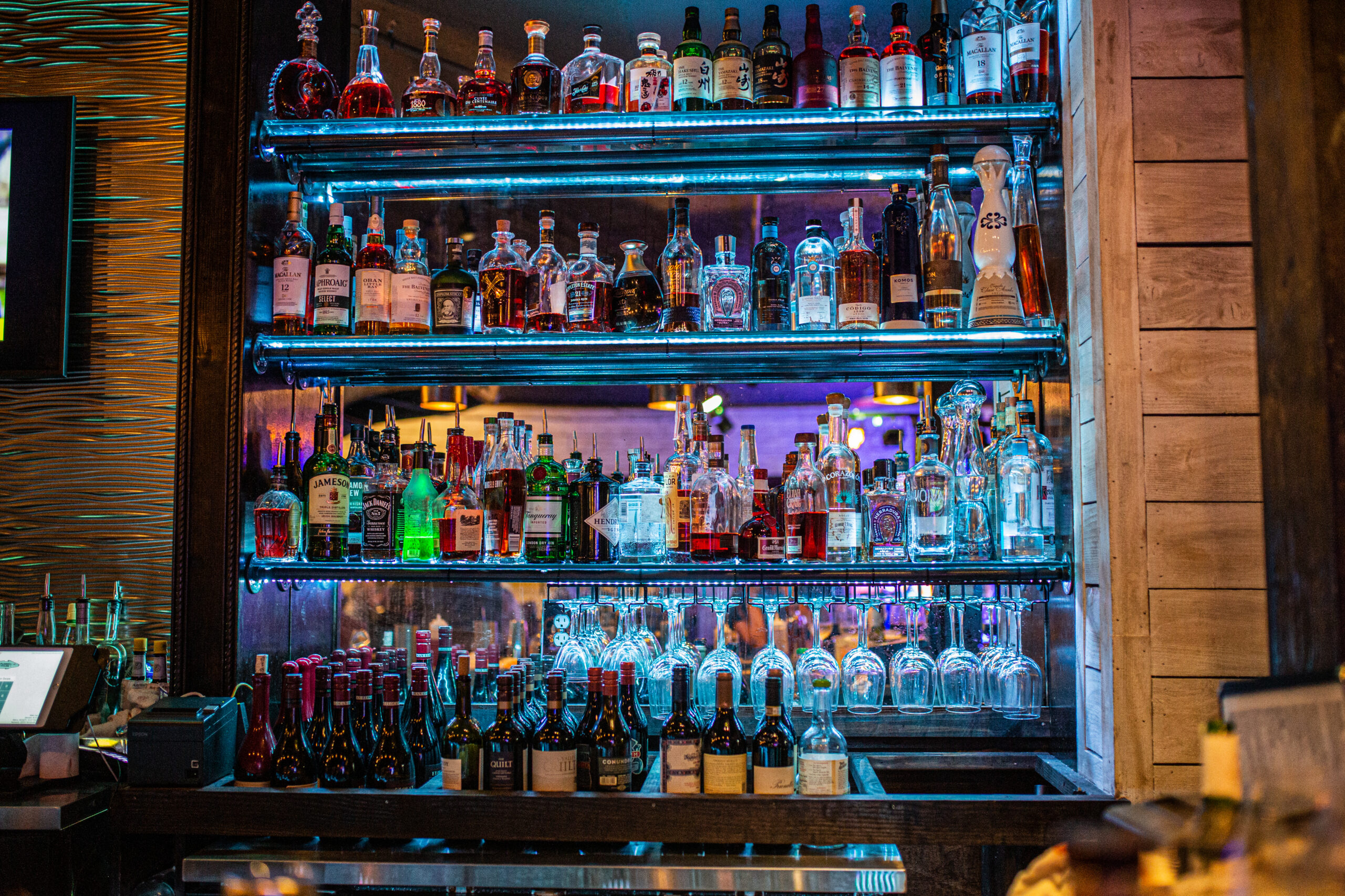 Backlit bar shelves displaying an extensive collection of premium spirits, wines, and hanging stemware, featuring whiskey, tequila, gin, rum, sake, and liqueurs arranged in a warm modern restaurant bar.